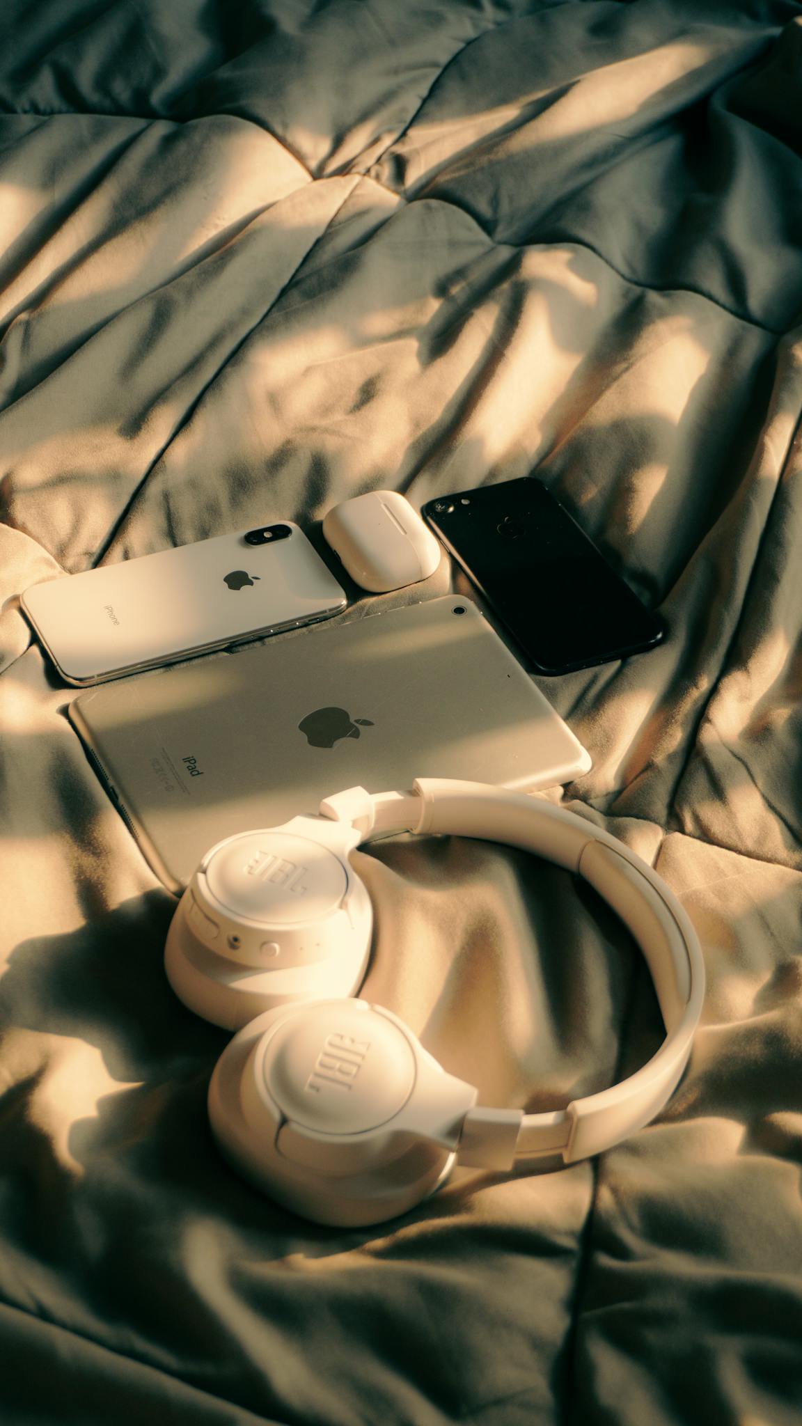 A collection of Apple devices including MacBook, iPad, and iPhone on a bed with sunlight.